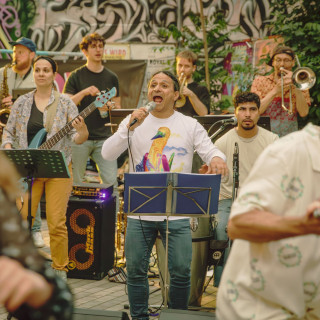 A band performs outdoors with a lead singer at a microphone, musicians playing various instruments, and graffiti art visible in the background.